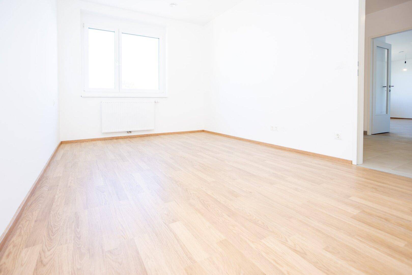 Helles Schlafzimmer mit Laminatboden und einem Fenster, das viel Tageslicht hereinlässt.
