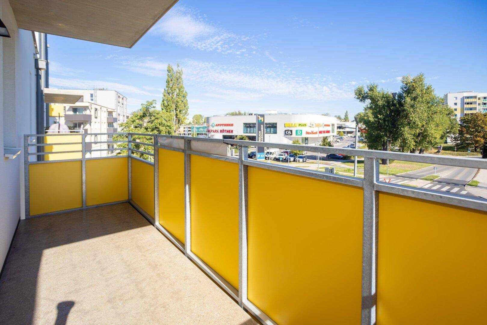 Balkon mit gelben Brüstungselementen und Ausblick auf eine belebte Straße und Geschäfte.