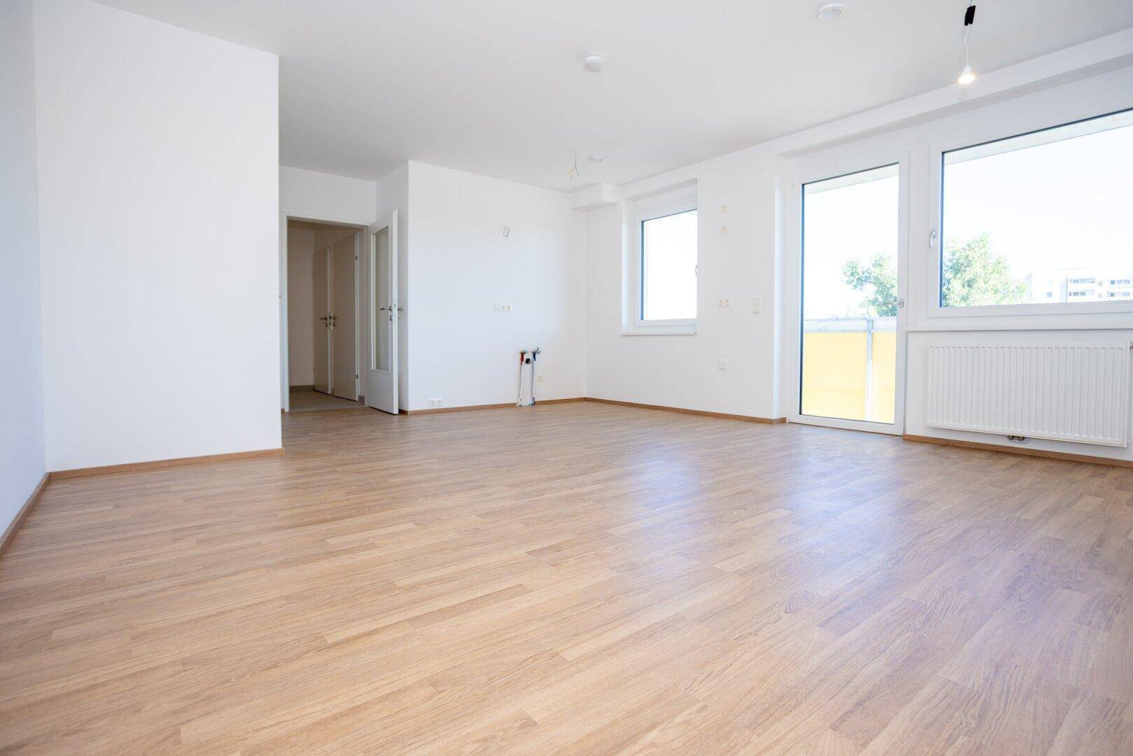 Geräumiges, helles Zimmer mit Laminatboden und großen Fenstern, ideal für ein Wohnzimmer.