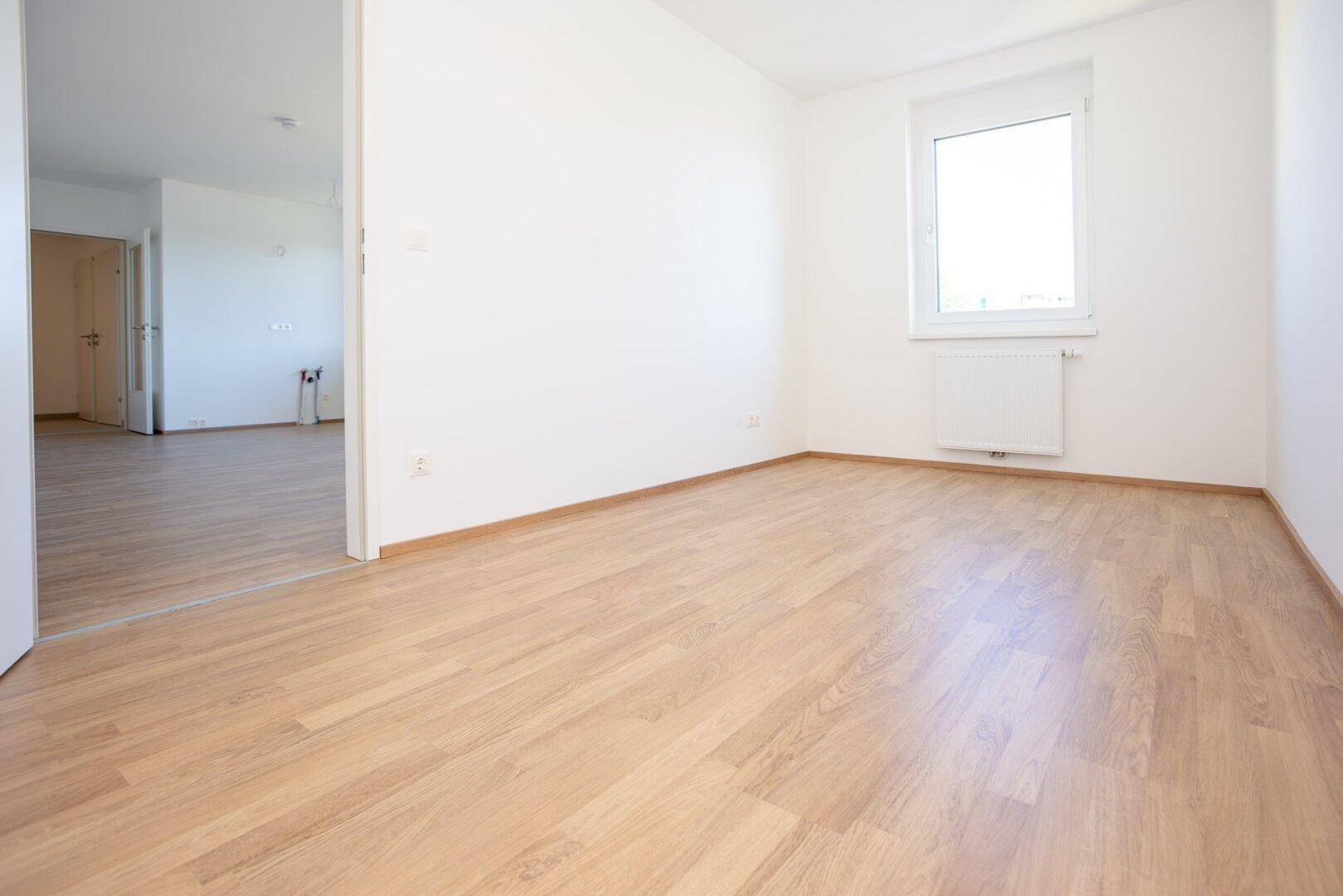 Helles Zimmer mit Laminatboden und offener Verbindung zu einem angrenzenden Bereich.
