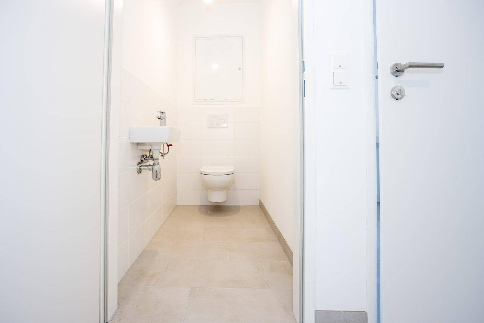 Kompaktes Gäste-WC mit wandhängender Toilette und kleinem Waschbecken, hell gefliest.
