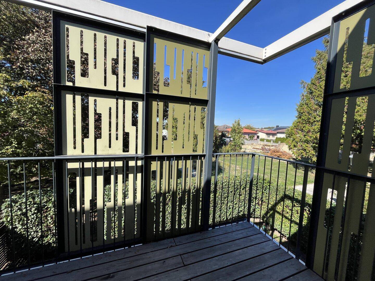 Sonniger Balkon mit Holzboden und modernen grünen Sichtschutzelementen, umgeben von Natur.