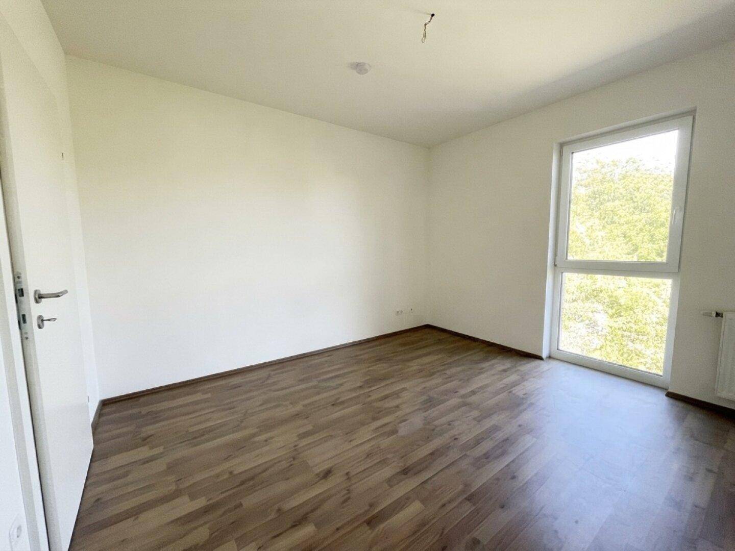 Helles Schlafzimmer mit Laminatboden und großem Fenster, das viel Tageslicht hereinlässt.