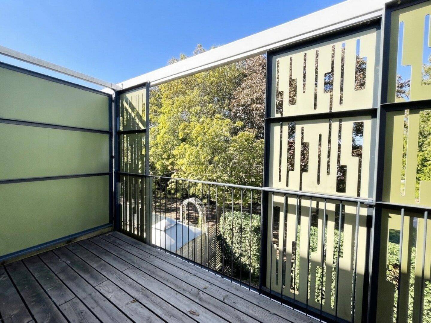 Moderner Balkon mit Holzdielen und dekorativen grünen Sichtschutzelementen, umgeben von Bäumen.