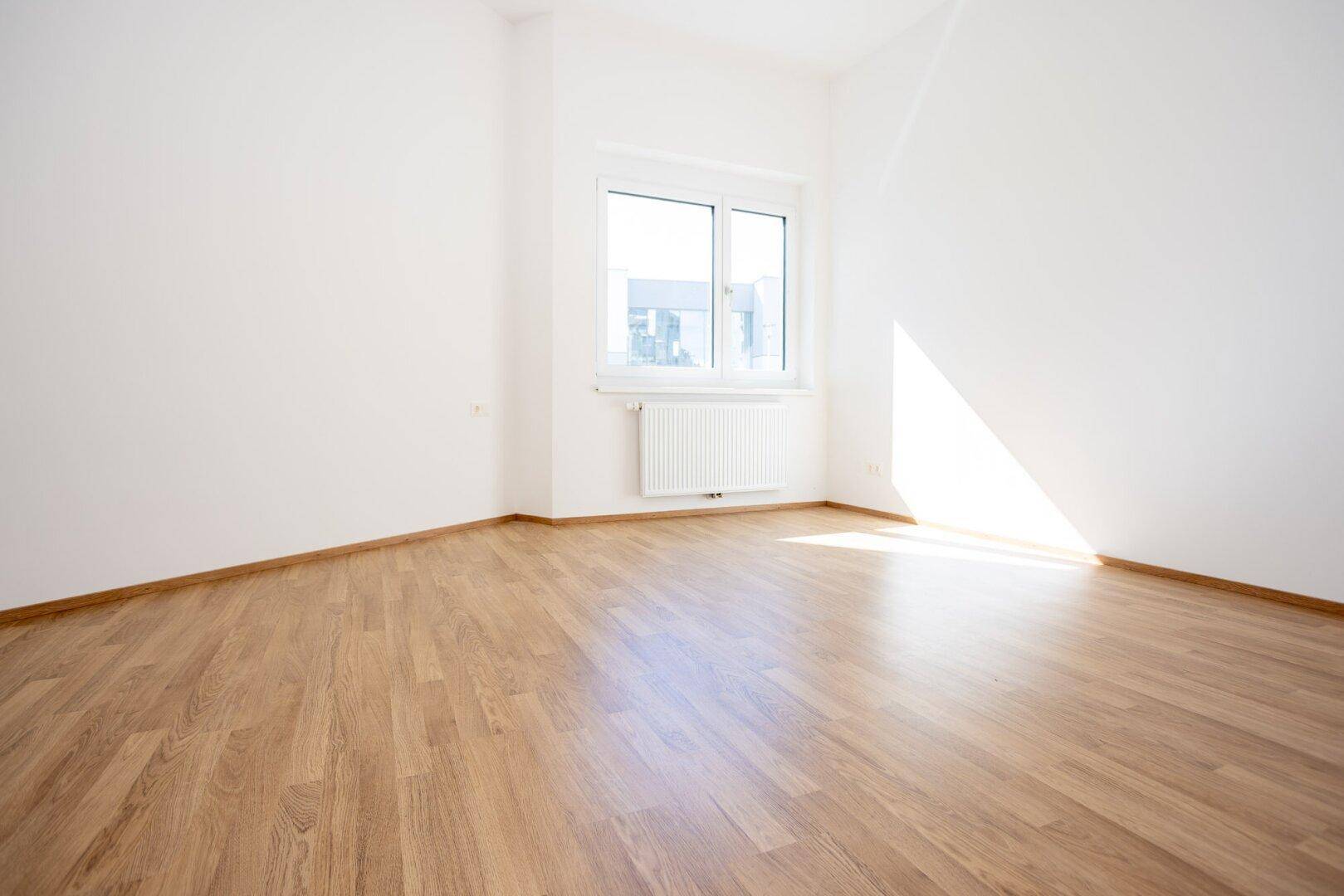 Sonniger, leerer Raum mit Laminatboden und großem Fenster, ideal als Schlaf- oder Arbeitszimmer.