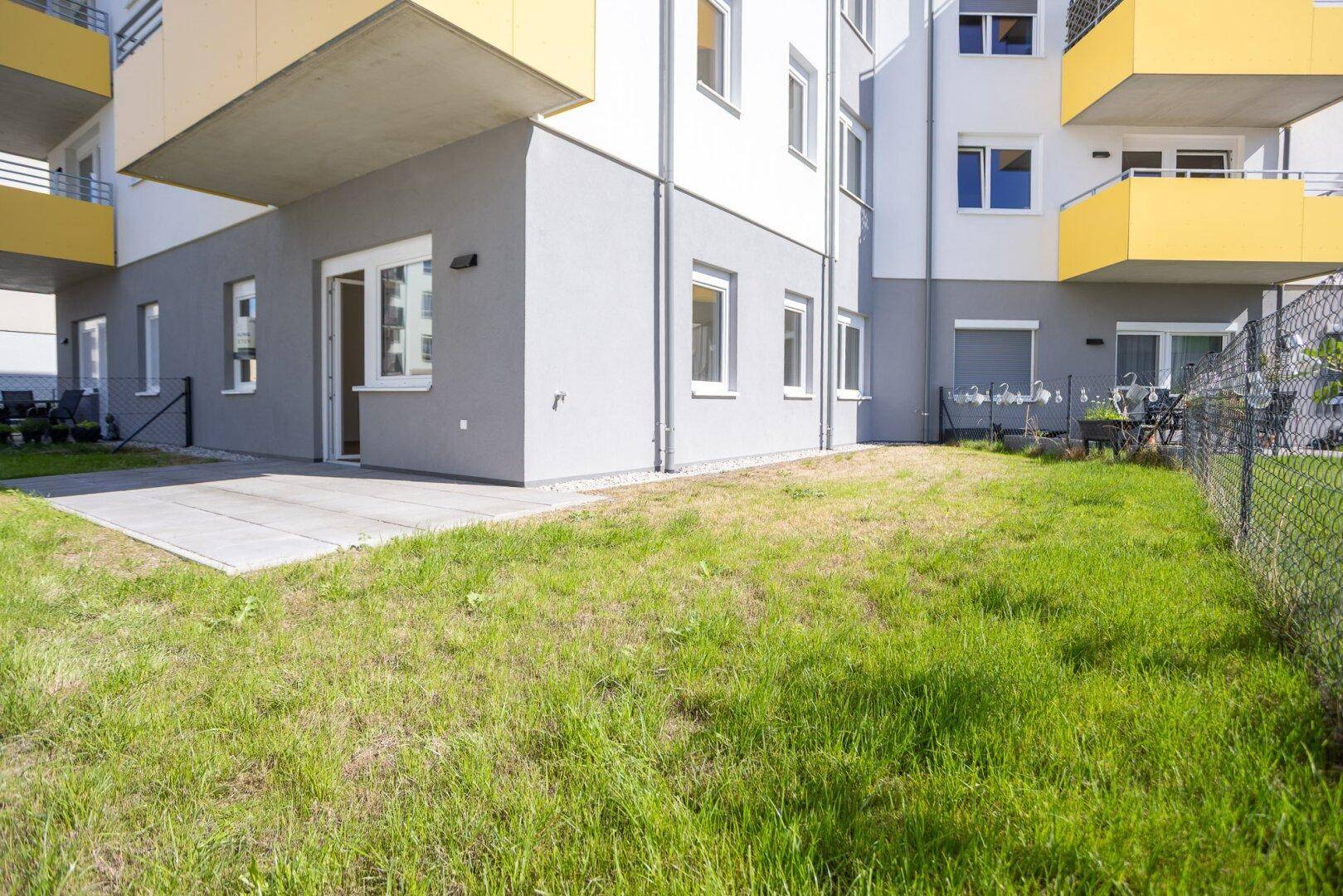 Weitläufiger Gartenbereich mit Terrasse und Blick auf die moderne Wohnanlage bei Sonnenschein.