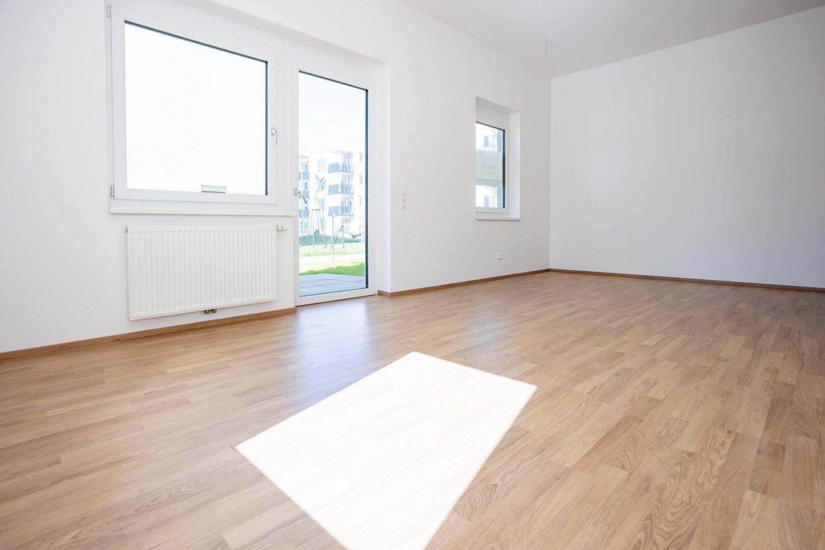 Helles, leeres Wohnzimmer mit Laminatboden und großem Fenster mit Zugang zum Außenbereich.