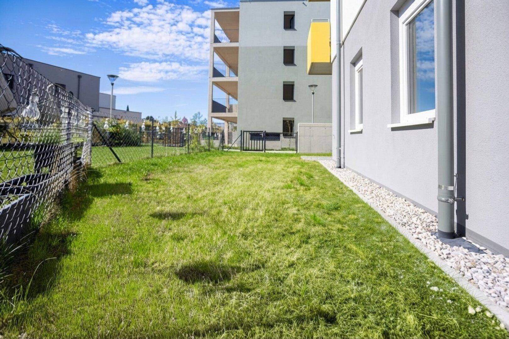 Grüner Rasenbereich entlang der Fassade eines modernen Wohngebäudes mit Kiesstreifen und Zaun.
