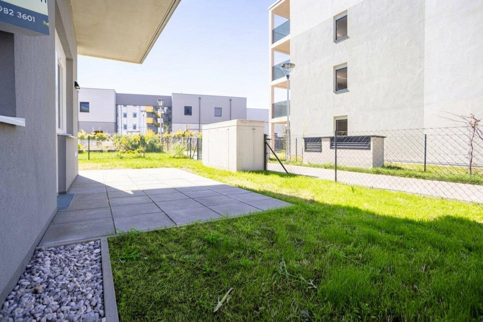 Sonniger Terrassenbereich mit angrenzendem Rasen und Kiesstreifen vor einem modernen Wohnhaus.