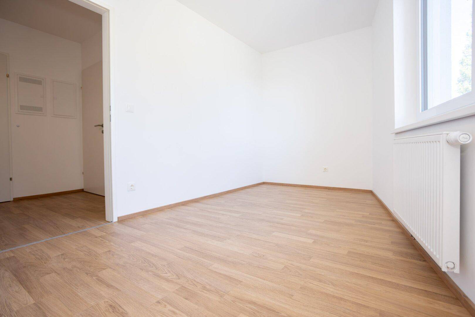 Leeres Zimmer mit Laminatboden und weiß gestrichenen Wänden, bereit zur Nutzung.