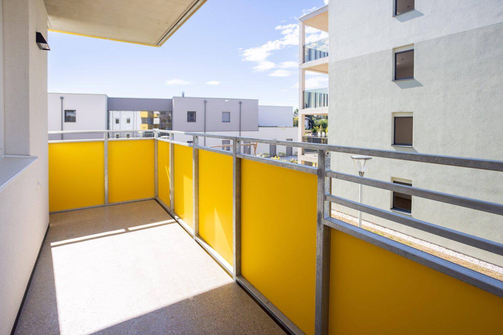 Sonniger Balkon mit gelben Paneelen und Ausblick auf die moderne Wohnanlage.