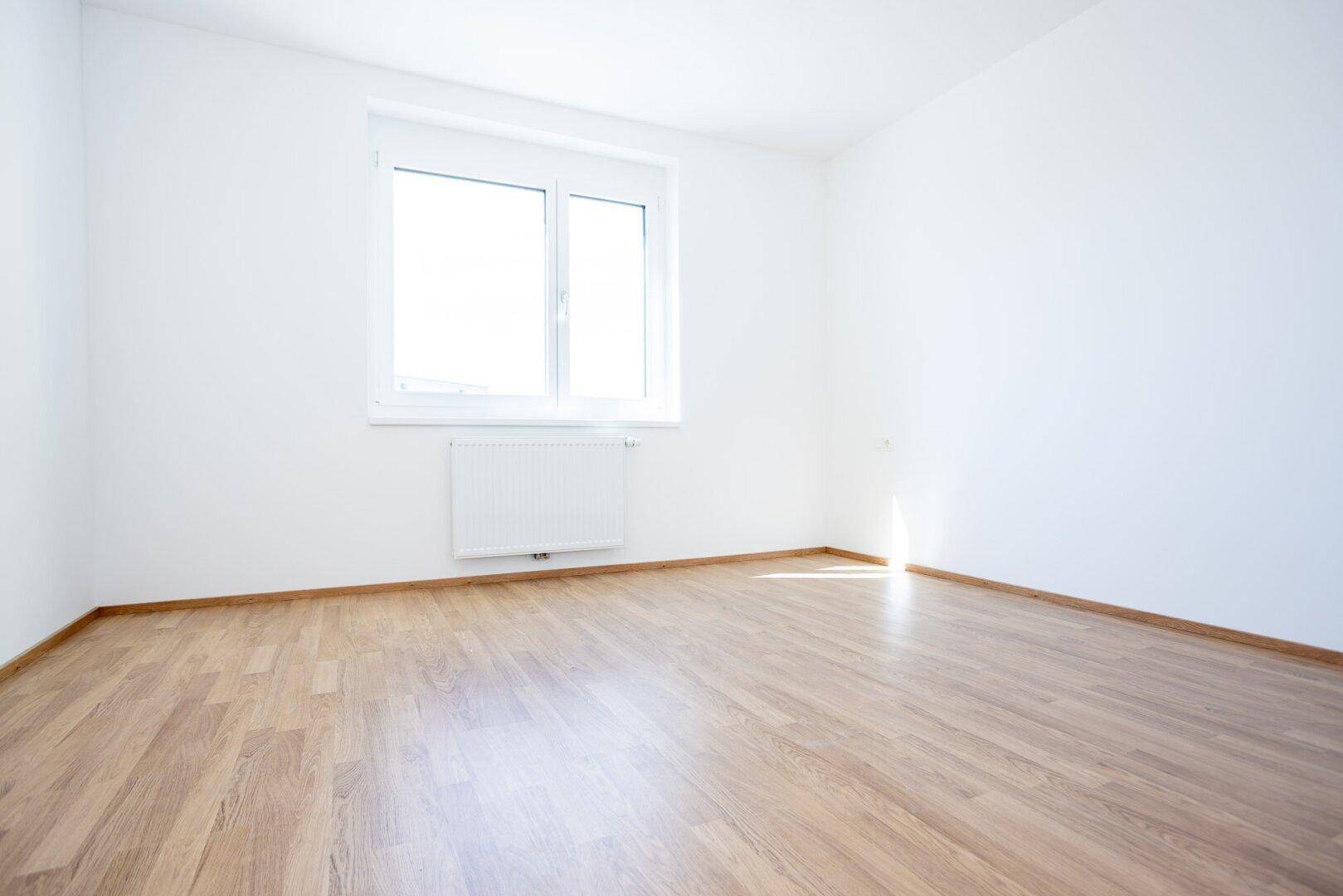 Helles Schlafzimmer mit Laminatboden, weißen Wänden und einem Fenster mit Heizkörper.