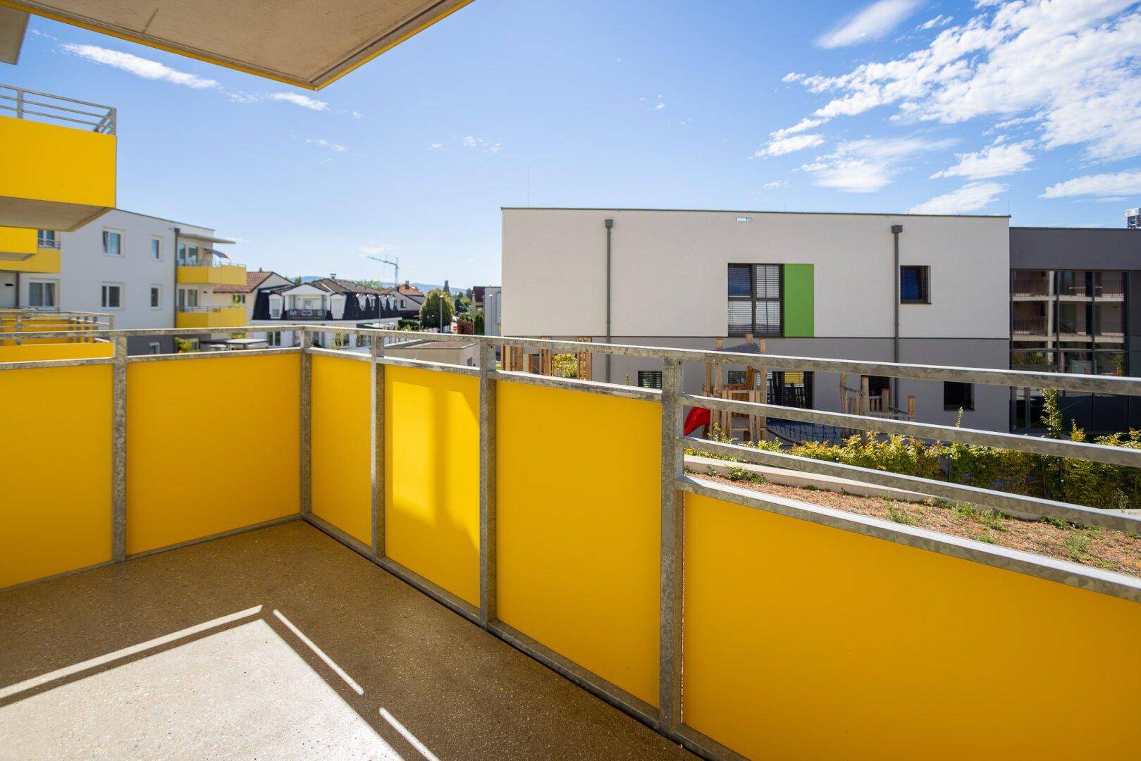 Sonniger Balkon mit gelben Brüstungen und Ausblick auf die umliegende Neubausiedlung.