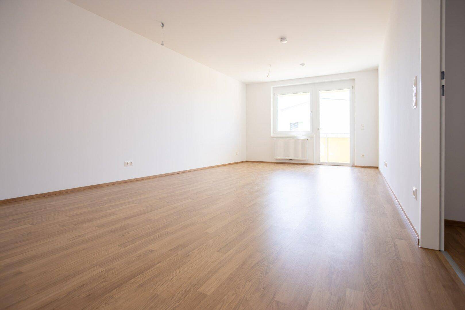 Helles, leeres Wohnzimmer mit Laminatboden und Zugang zu einem Balkon oder einer Terrasse.