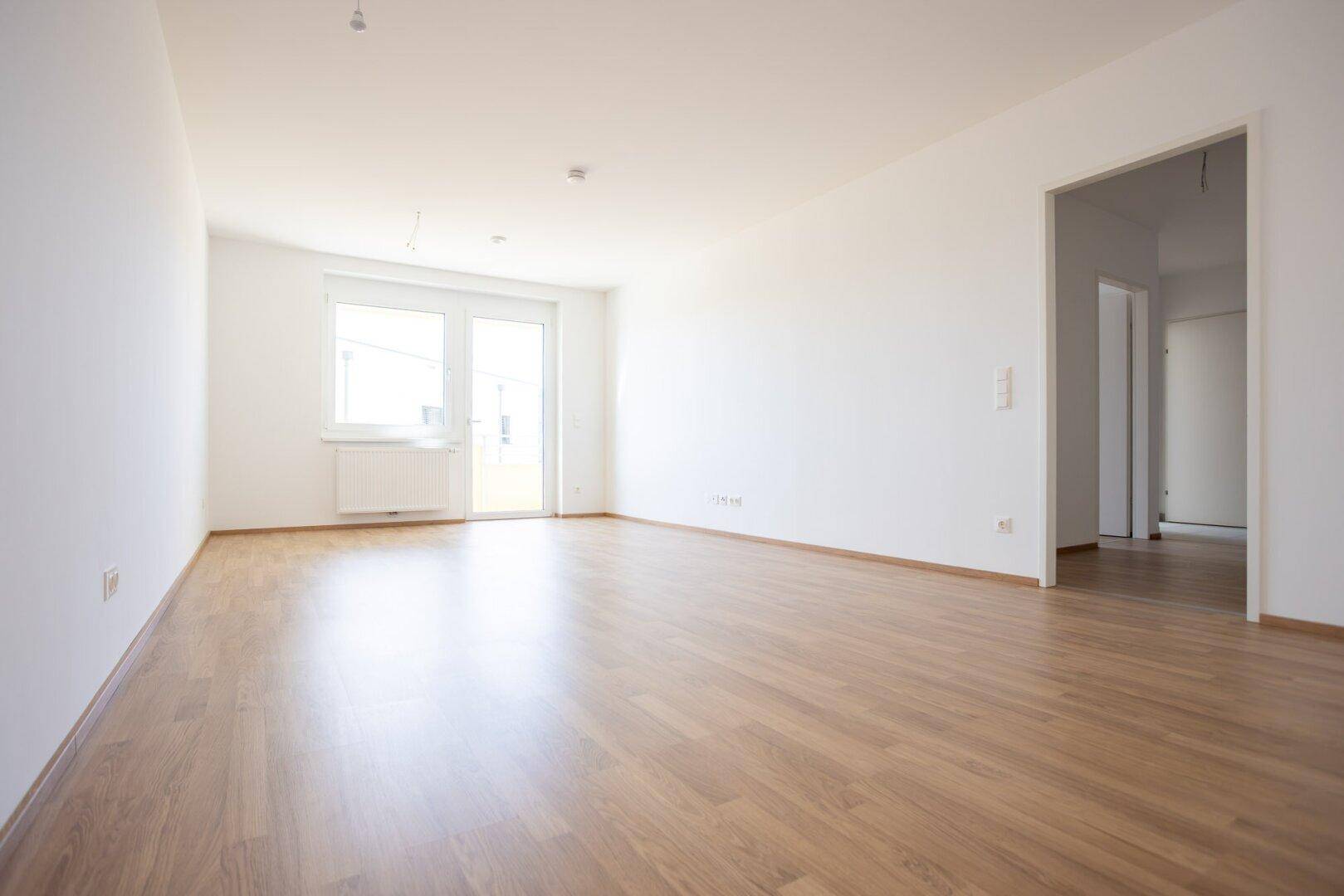 Geräumiges, unmöbliertes Wohnzimmer mit hellem Laminatboden und Blick auf den Flur.