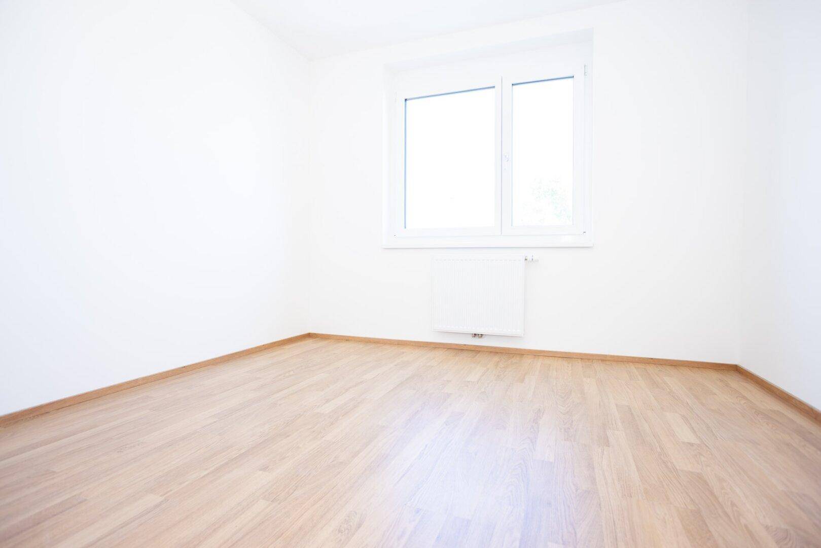 Helles, leeres Zimmer mit Laminatboden und einem großen Fenster, ideal als Schlafzimmer.