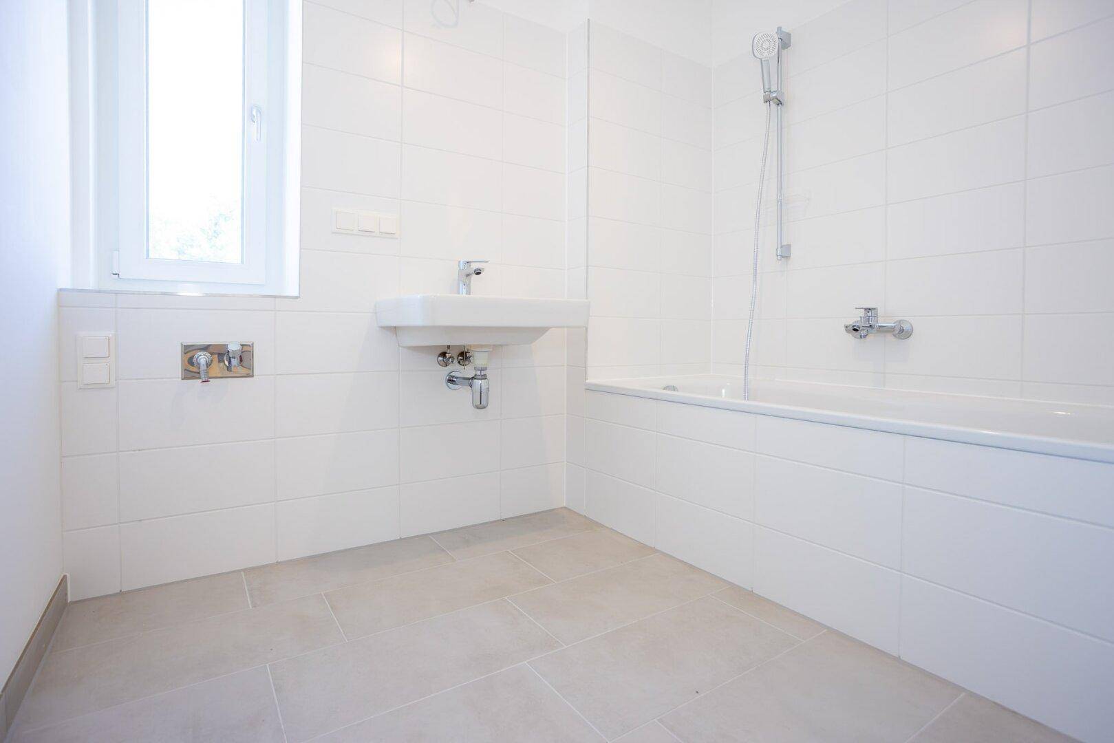 Modernes Badezimmer mit weißen Fliesen, Badewanne, Waschbecken und Fenster für Tageslicht.