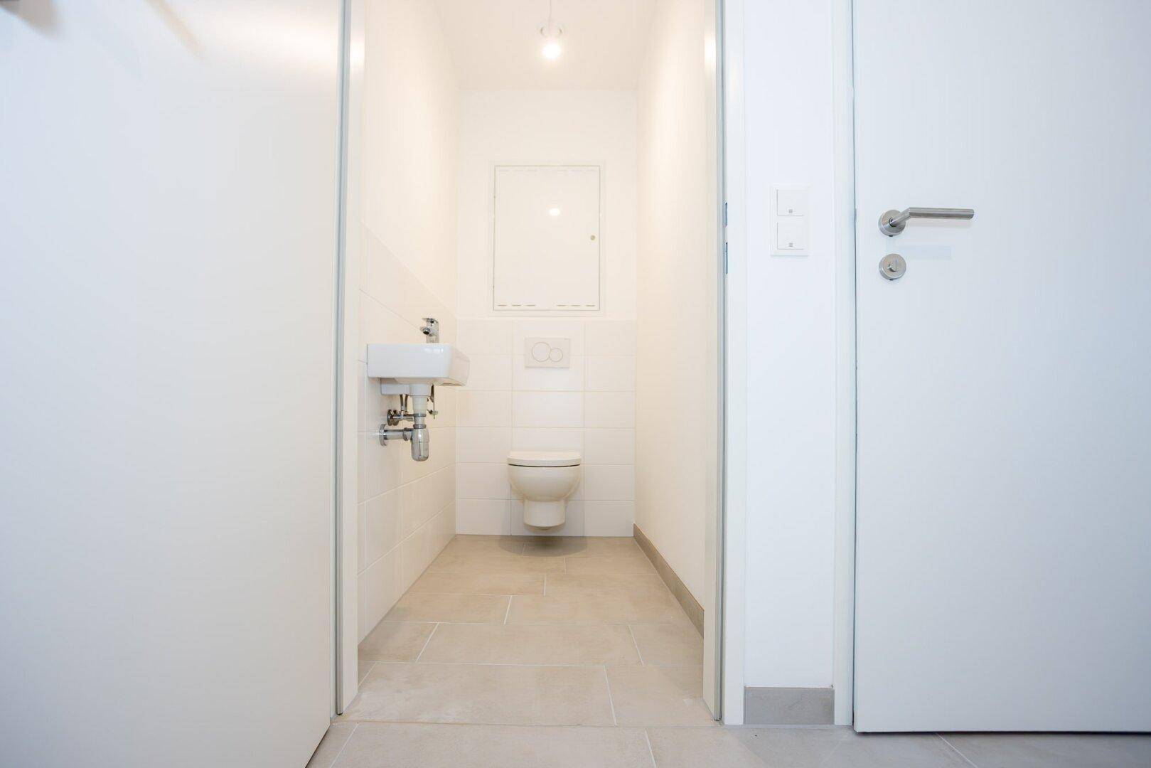 Separates Gäste-WC mit wandhängender Toilette, kleinem Waschbecken und hellen Fliesen.
