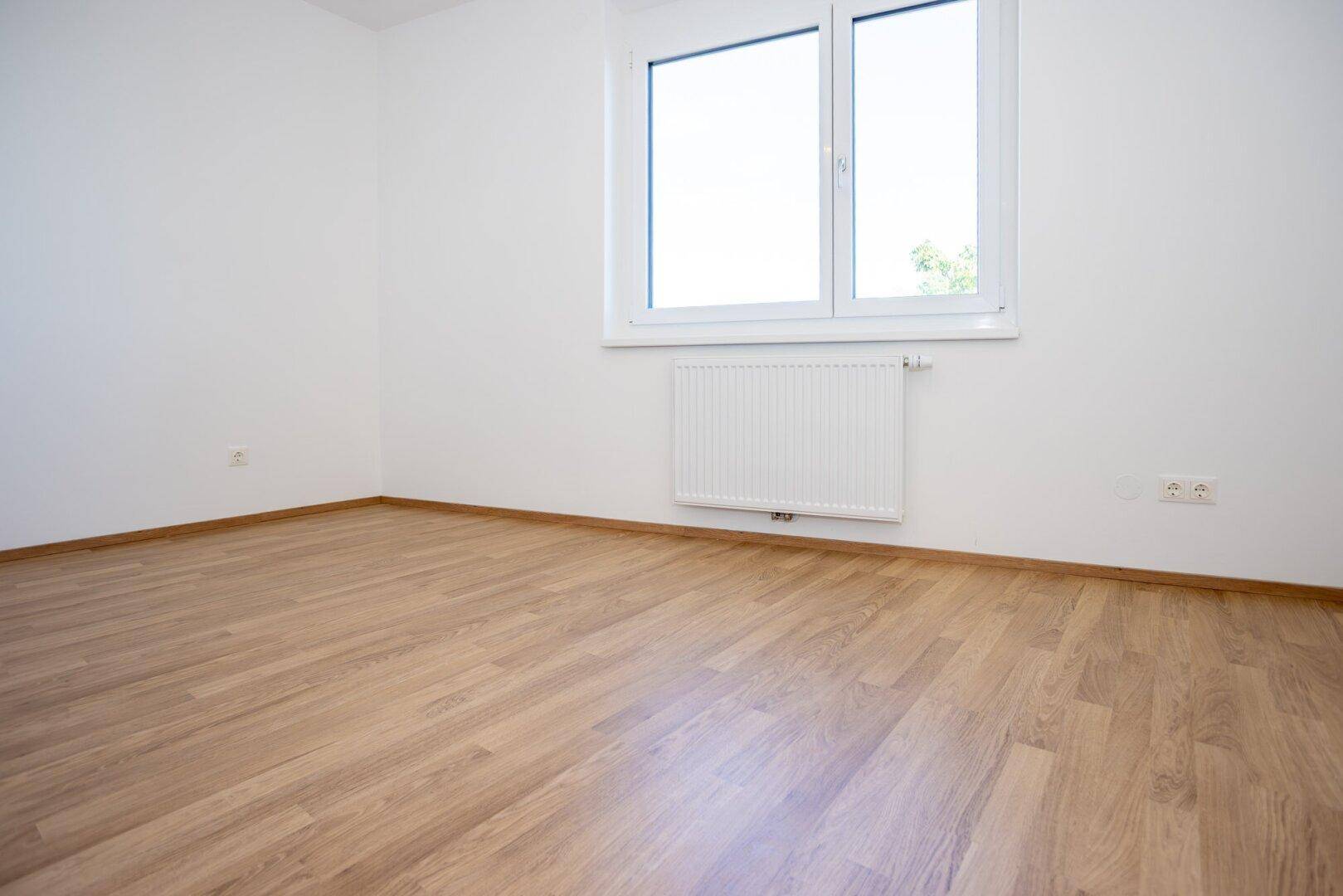 Helles Schlafzimmer mit Laminatboden, großem Fenster und Heizkörper.