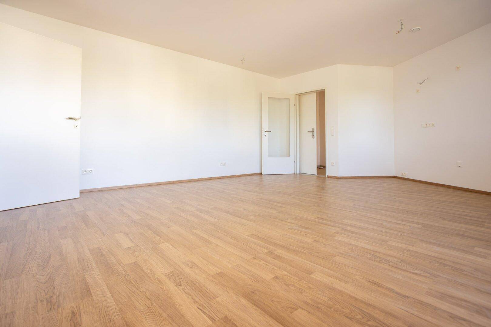 Geräumiges, leeres Zimmer mit Laminatboden und zwei Türen.