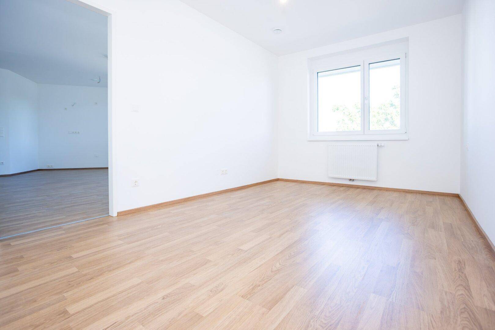 Heller Wohnbereich mit Laminatboden und offenem Übergang zu einem weiteren Zimmer.