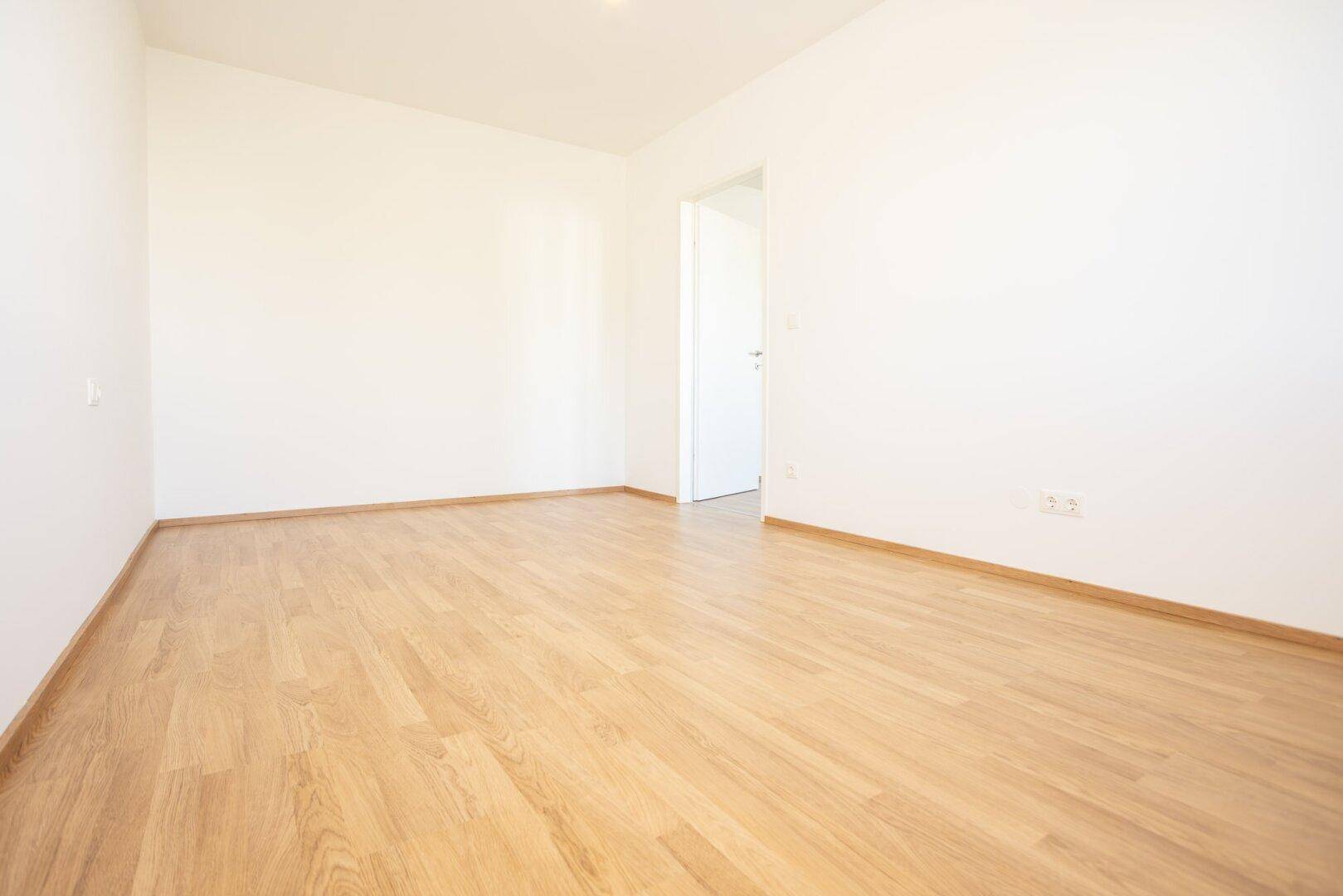 Leeres Zimmer mit Laminatboden, weiß gestrichenen Wänden und Türzugang.