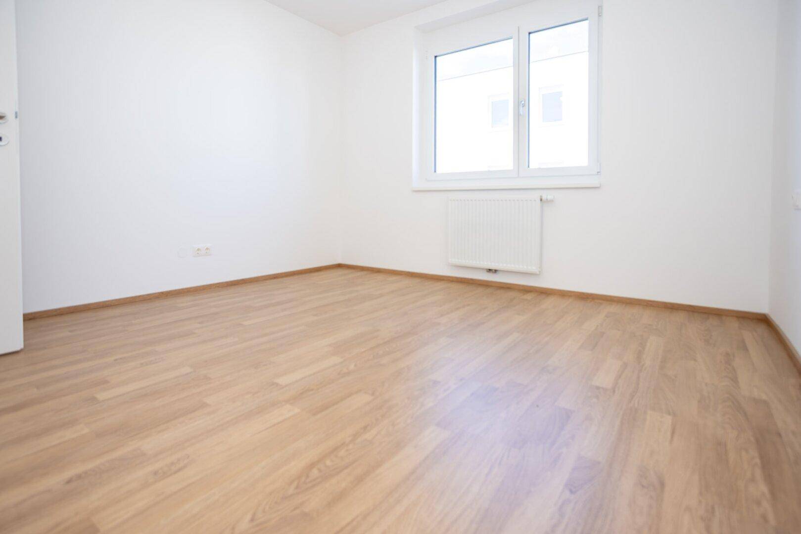 Helles Schlafzimmer mit Laminatboden, weißen Wänden und einem großen Fenster.