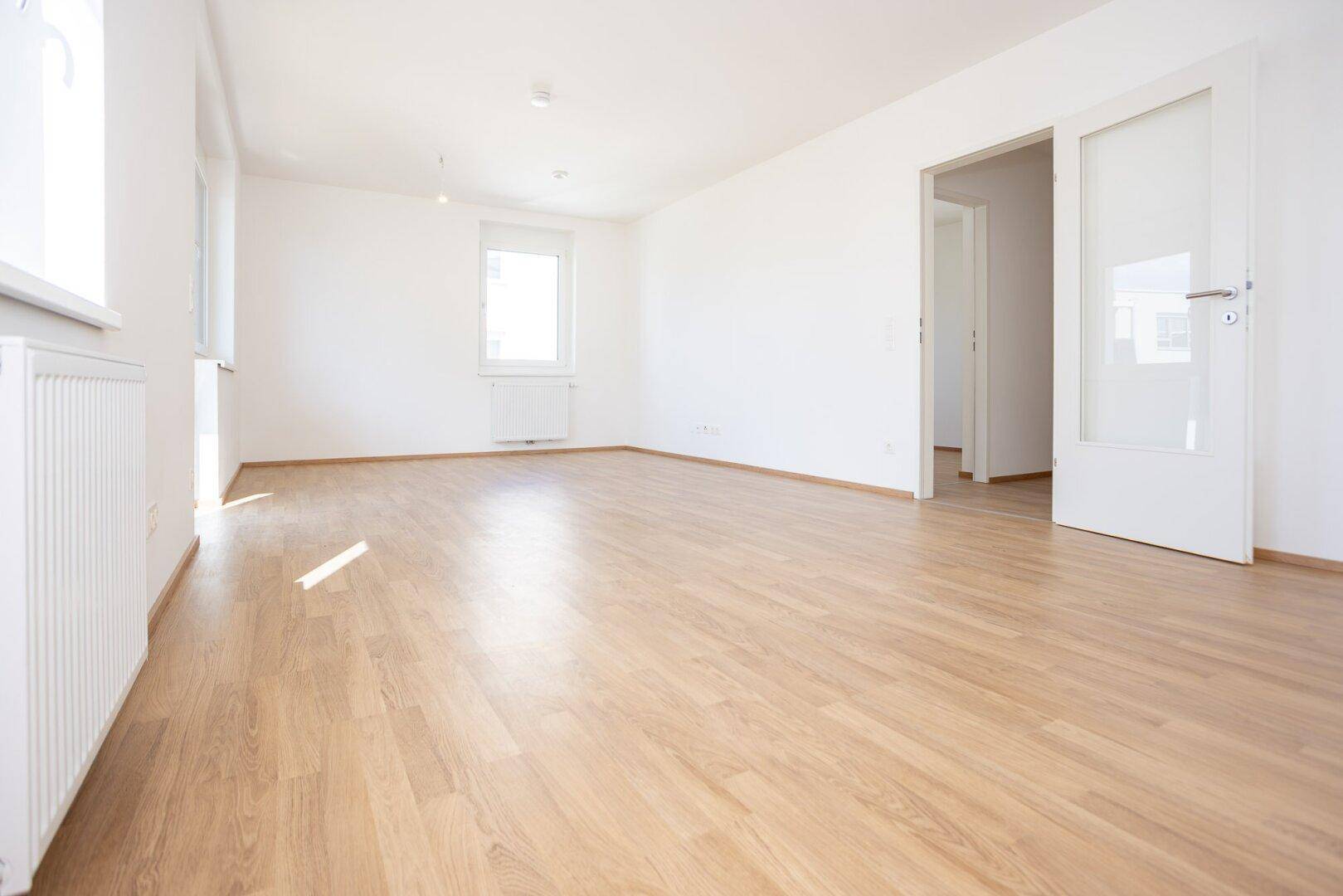 Geräumiges, helles Zimmer mit Laminatboden, weißen Wänden und einer Tür zu weiteren Räumen.
