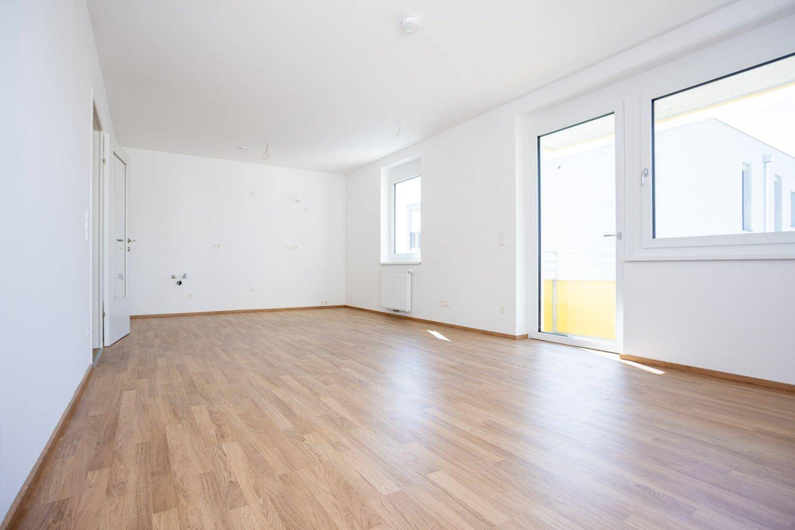 Lichtdurchfluteter Raum mit hellem Laminatboden, weißen Wänden und direktem Zugang zum Balkon.