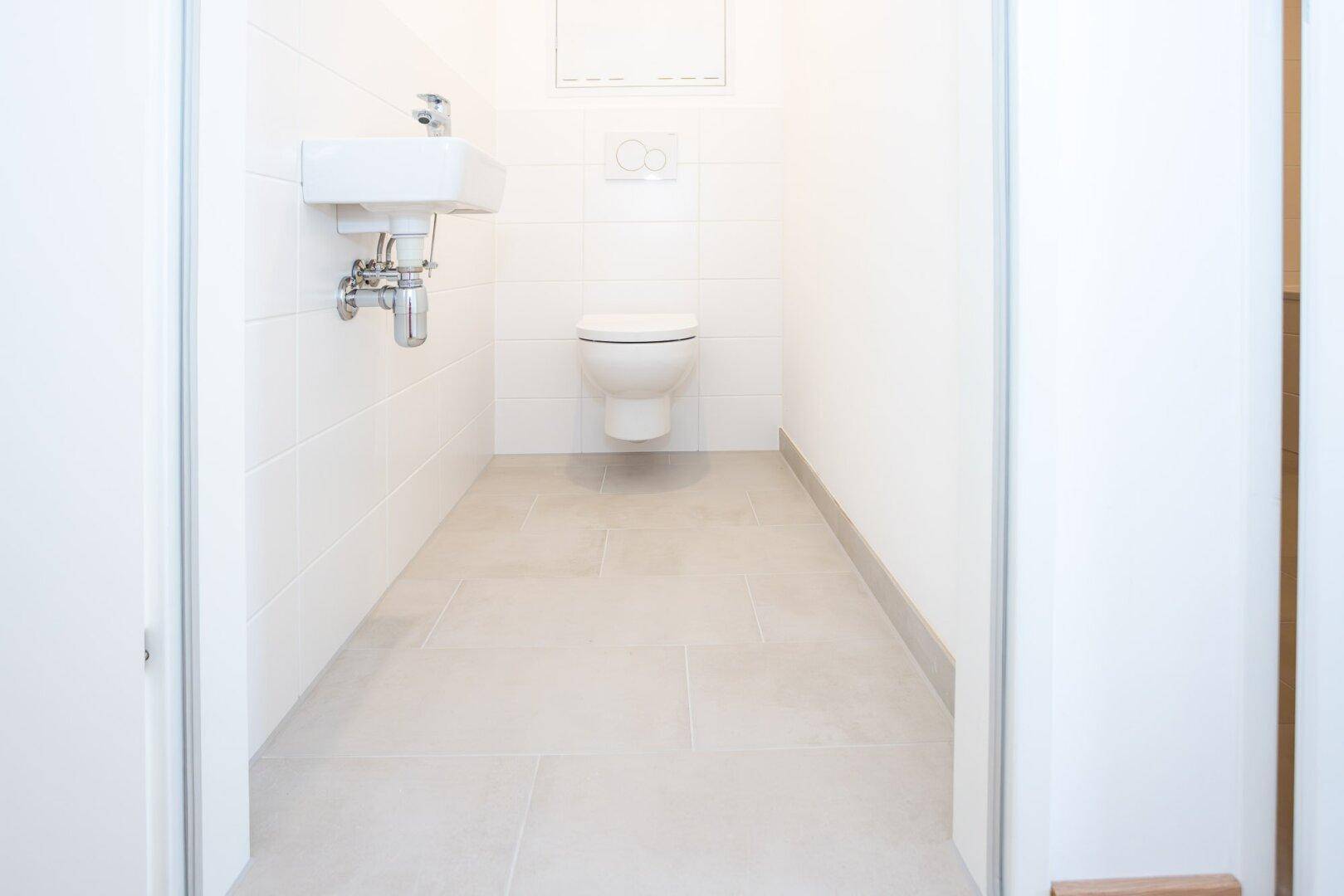 Separates Gäste-WC mit wandhängender Toilette, kleinem Waschbecken und hellen Fliesen.