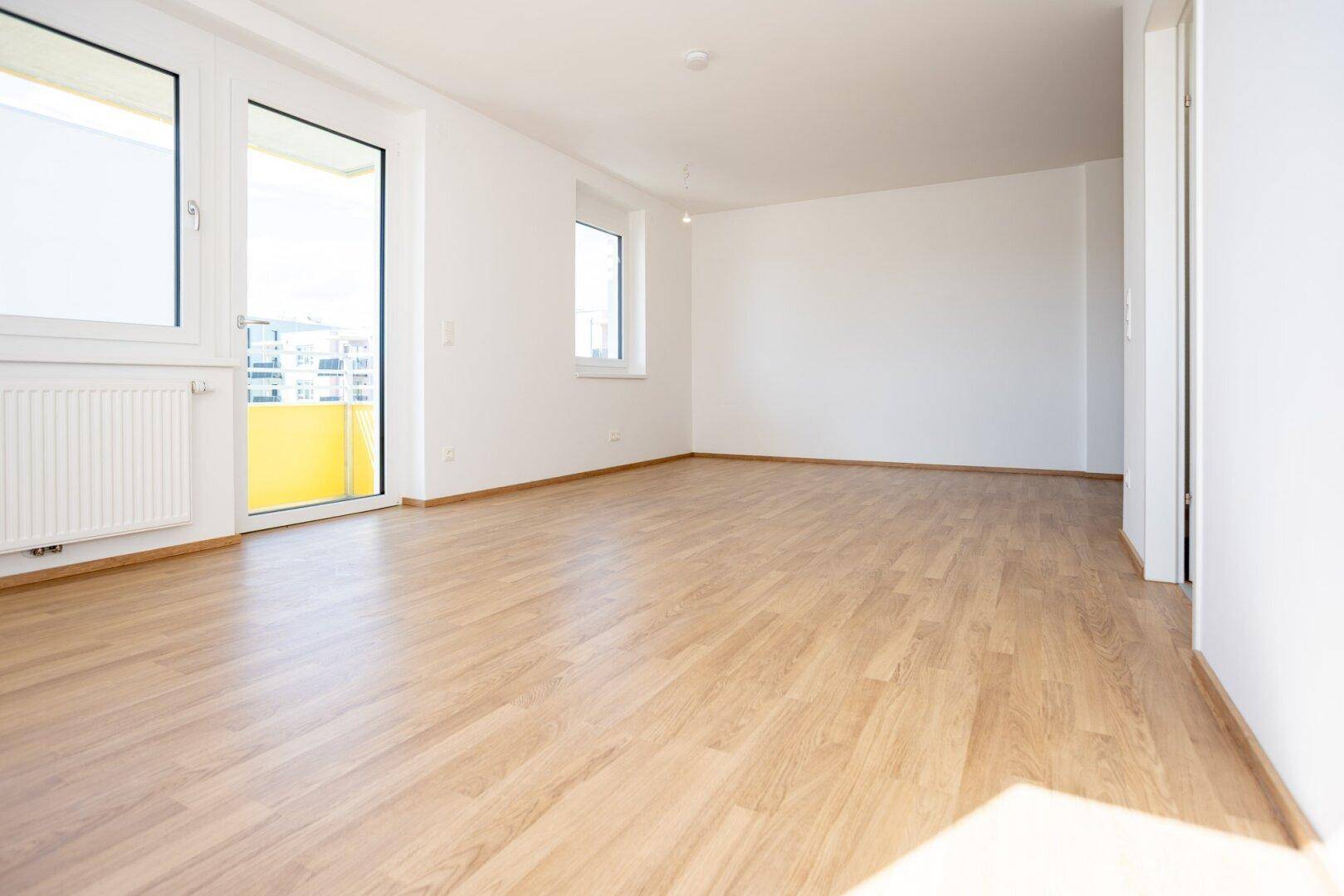Helles Wohnzimmer mit modernem Laminatboden, weißen Wänden und Zugang zu einem sonnigen Balkon.