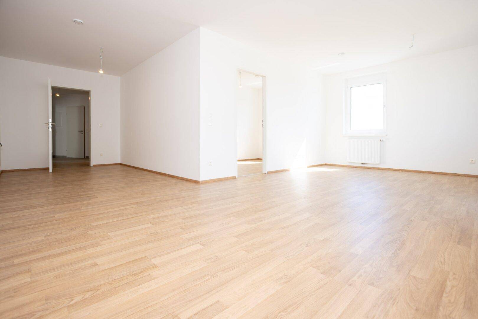 Lichtdurchfluteter Raum mit hellem Laminatboden, weißen Wänden und mehreren Türöffnungen.