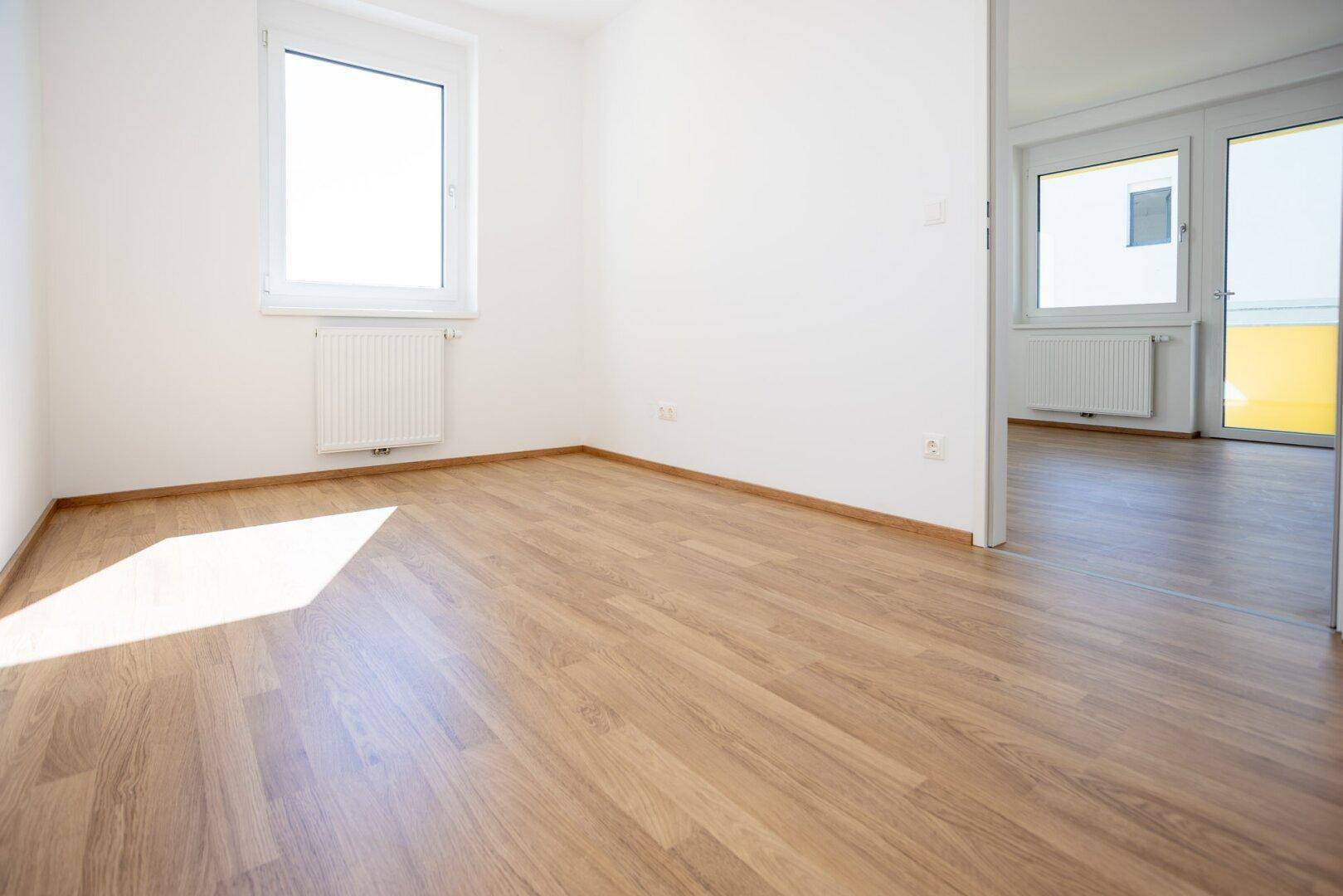 Helles Zimmer mit Laminatboden, weißen Wänden und Zugang zu einem weiteren Raum mit Balkon.