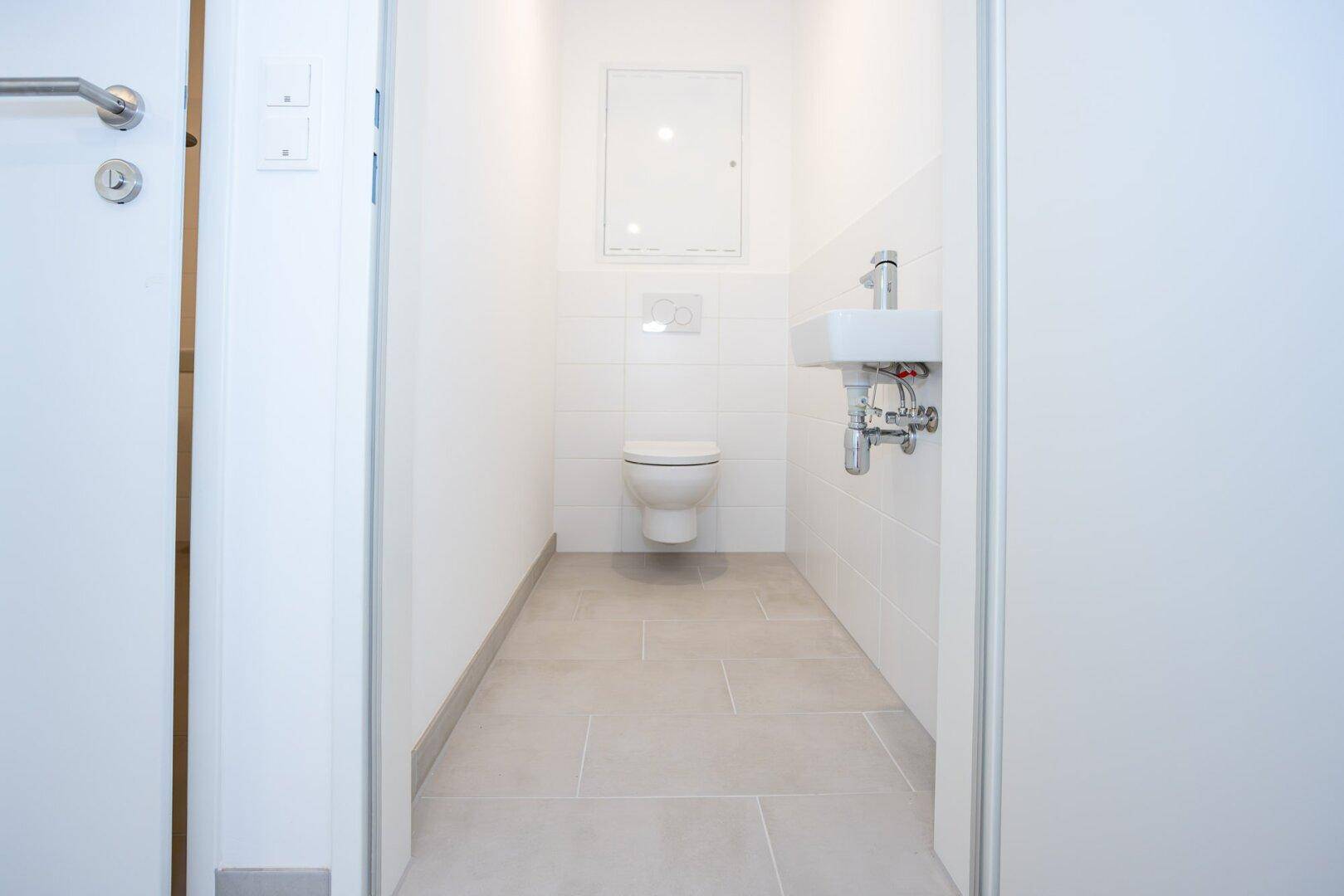Separates Gäste-WC mit wandhängender Toilette, kleinem Waschbecken und hellen Fliesen.