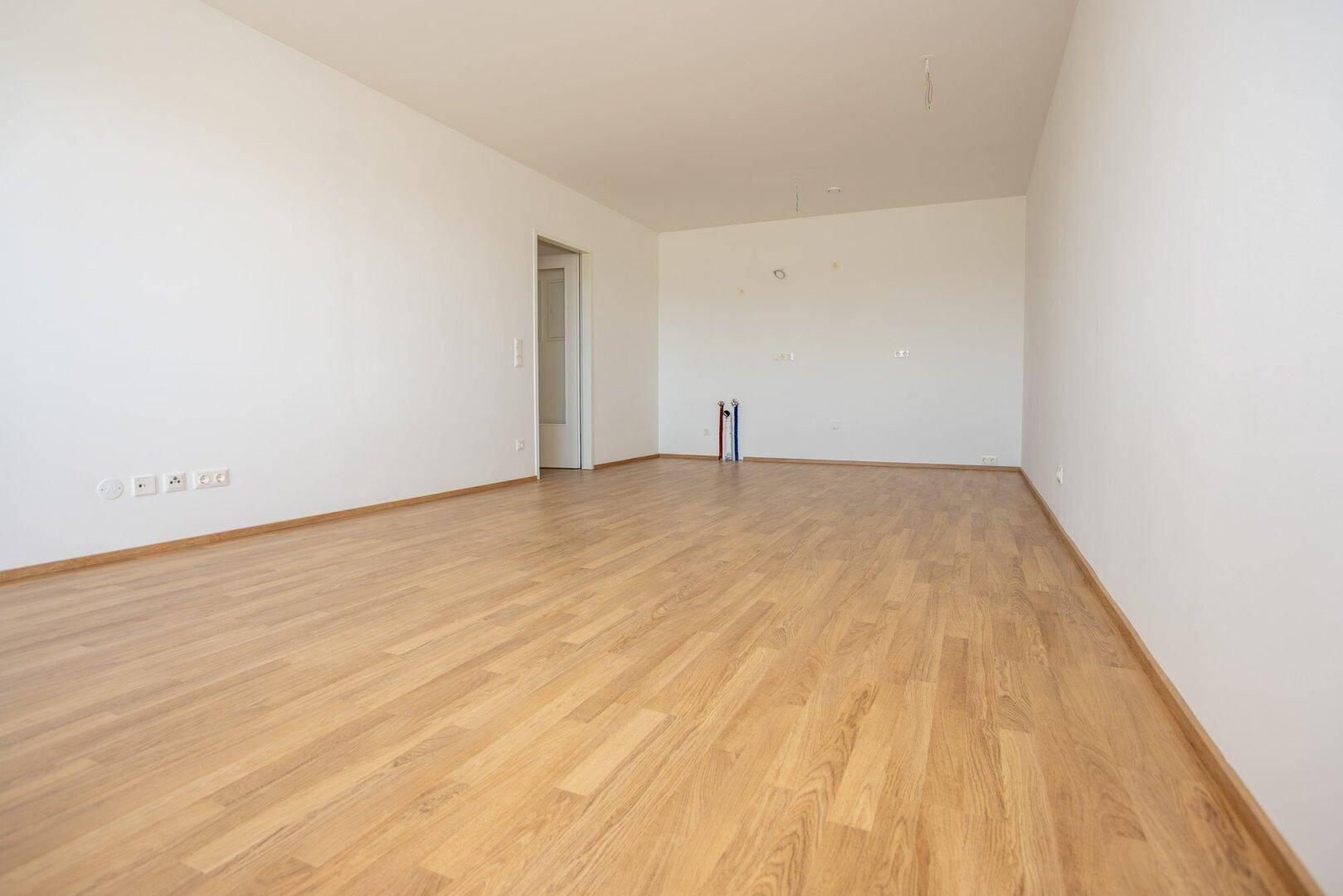 Leerer Küchenraum mit hellem Laminatboden, weißen Wänden und vorbereiteten Anschlüssen für eine Einbauküche.