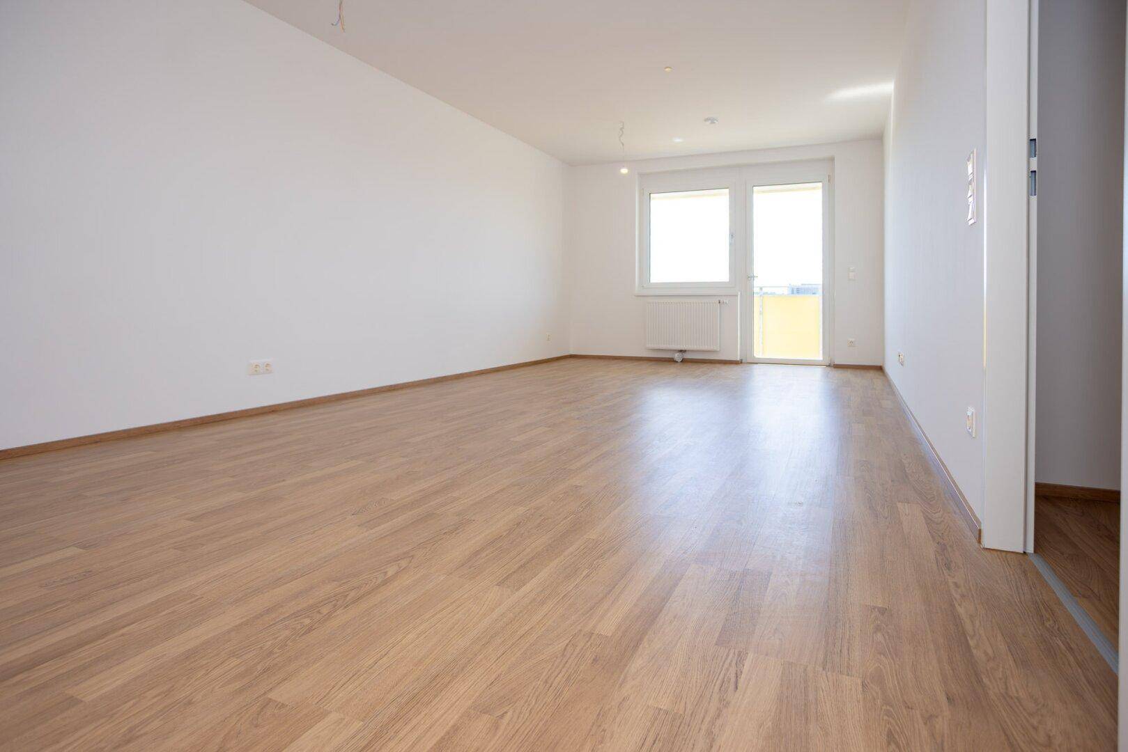 Helles, leeres Wohnzimmer mit Laminatboden, weißen Wänden und Zugang zu einem Balkon.