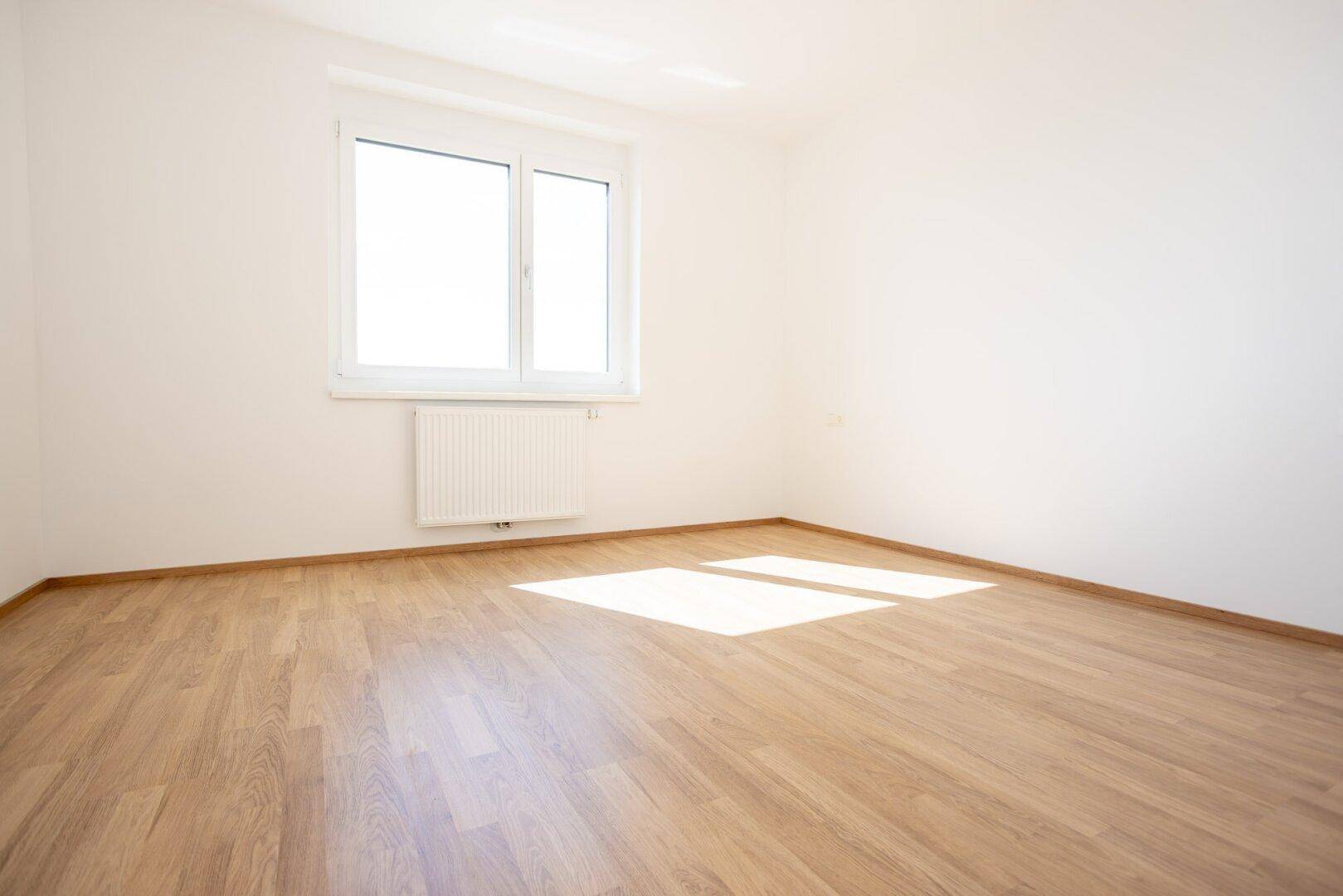 Helles, leeres Schlafzimmer mit Laminatboden, weißen Wänden und viel natürlichem Tageslicht.