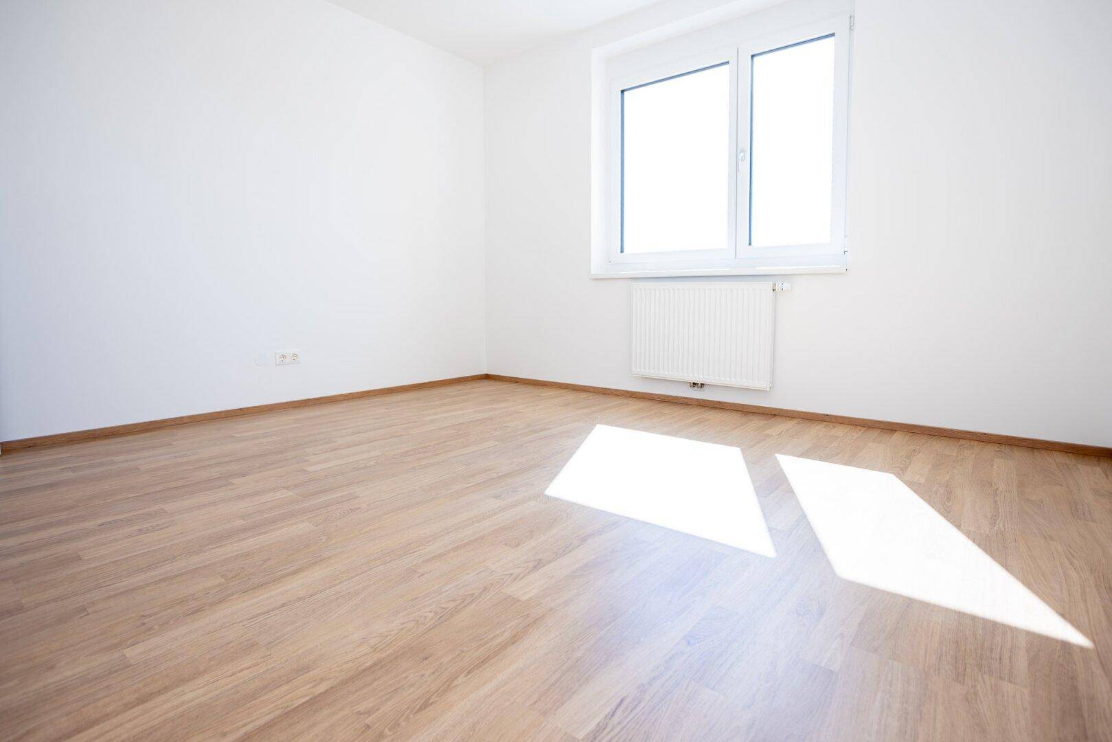 Freundliches, unmöbliertes Zimmer mit Laminatboden und einem Fenster, das viel Sonnenlicht hereinlässt.