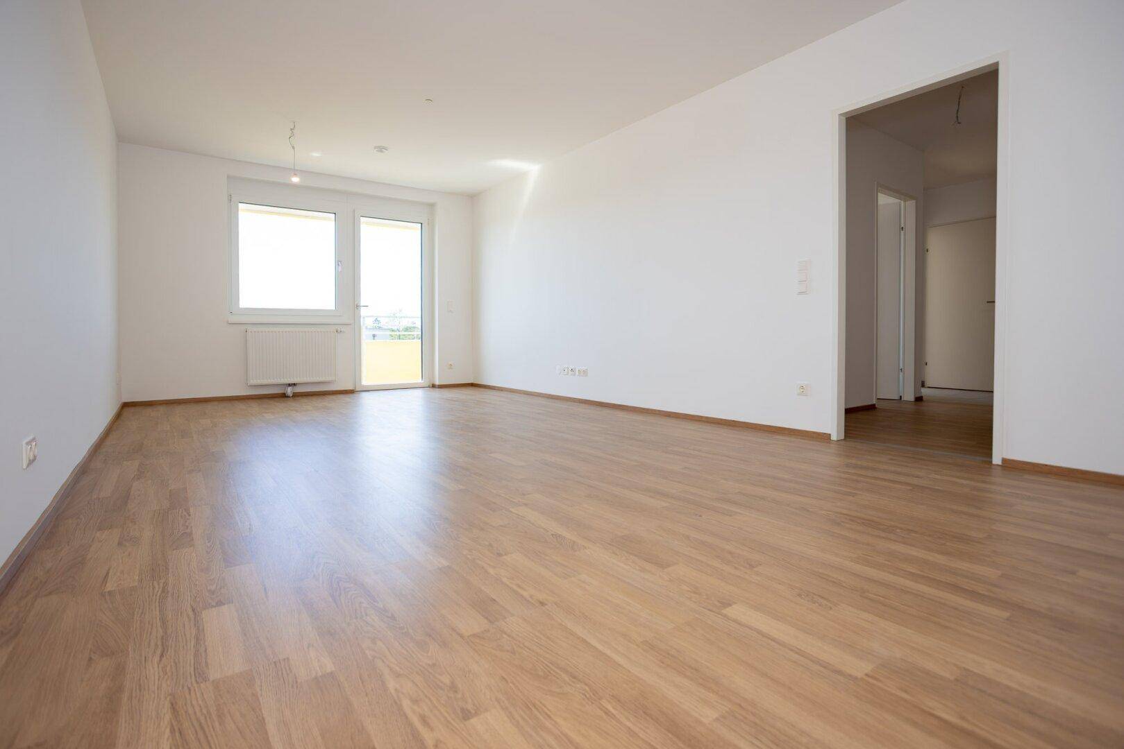 Geräumiges, unmöbliertes Wohnzimmer mit hellem Laminatboden und offenem Zugang zu weiteren Räumen.