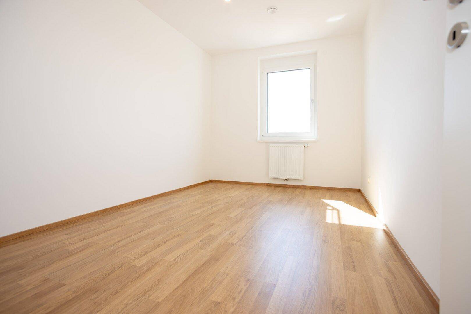 Leeres Zimmer mit hellem Laminatboden, weißen Wänden und einem Fenster, ideal als Schlaf- oder Arbeitszimmer.