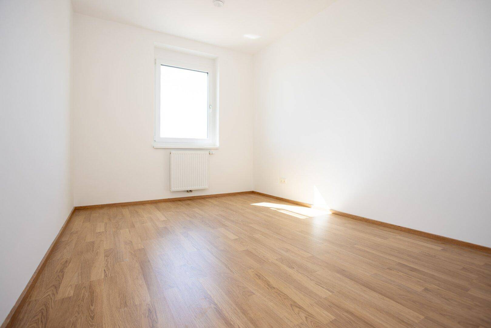 Unmöbliertes Zimmer mit Laminatboden und weißen Wänden, das durch das Fenster gut belichtet wird.