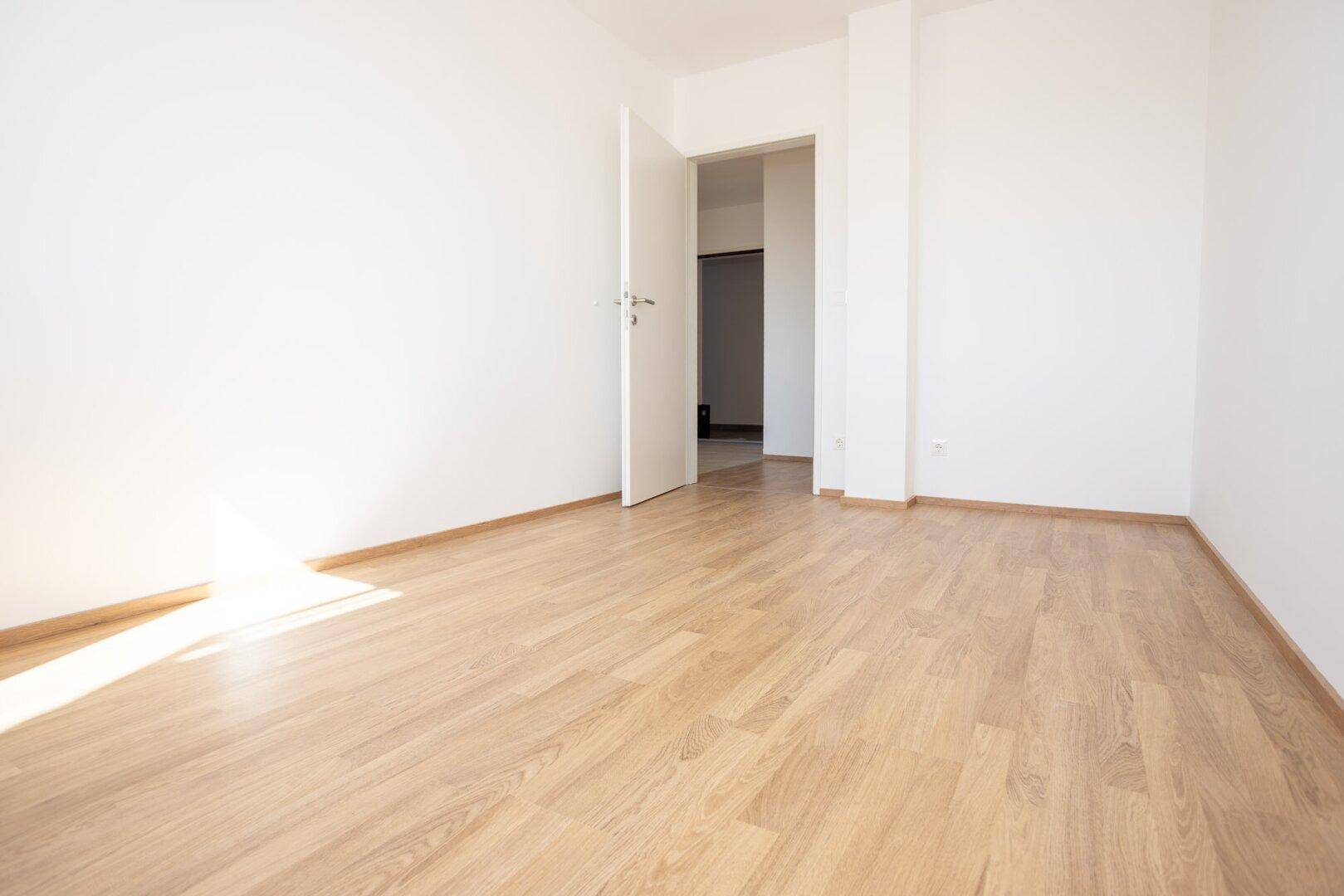Leerer Raum mit Laminatboden und weißen Wänden, der durch eine offene Tür mit einem weiteren Bereich verbunden ist.