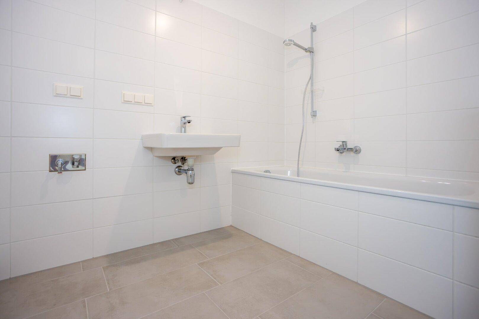 Modernes Badezimmer mit weißen Wandfliesen, grauen Bodenfliesen, Badewanne mit Duschfunktion und Waschbecken.
