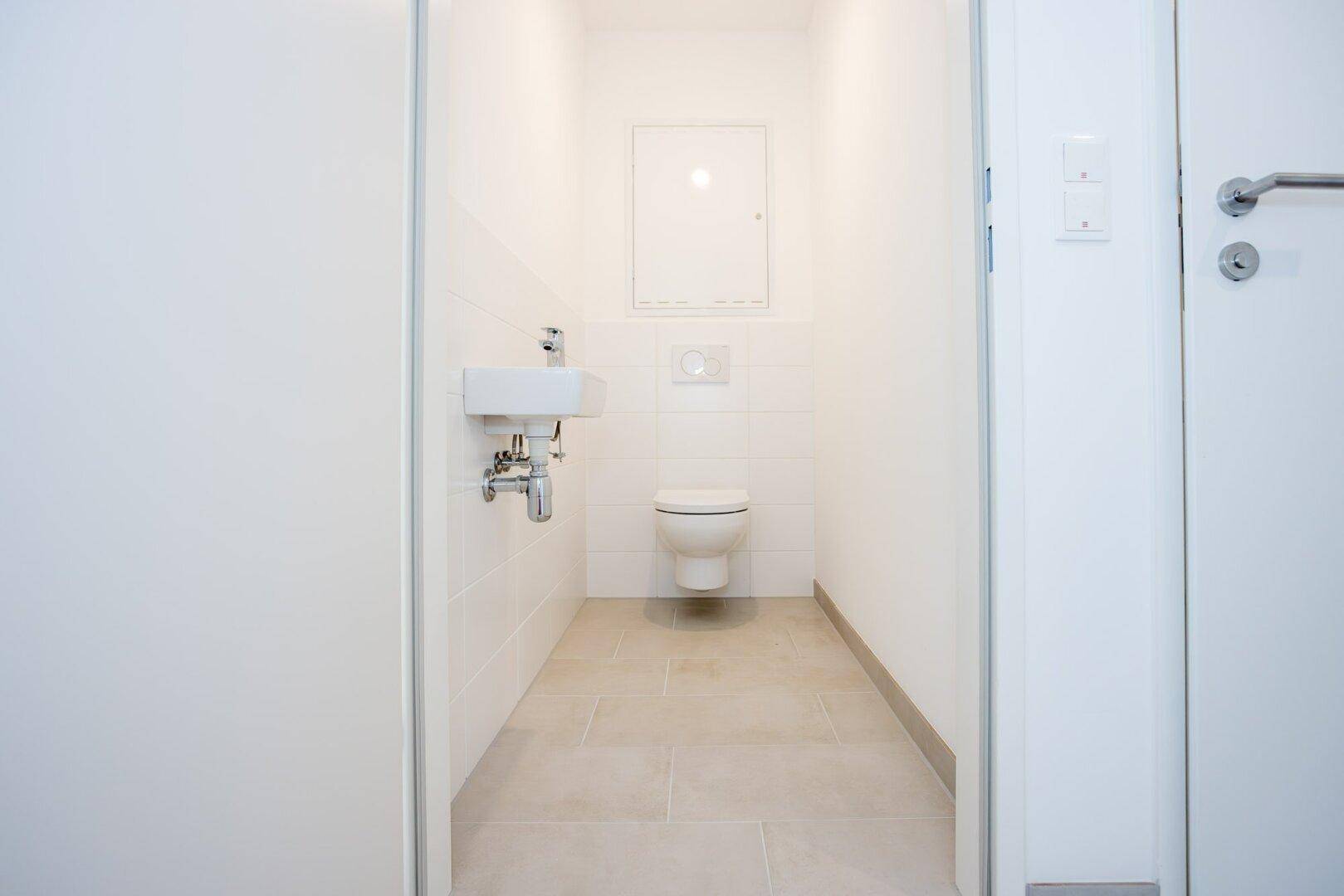 Kleines Gäste-WC mit weißen Wandfliesen, grauen Bodenfliesen, wandhängendem WC und Handwaschbecken.