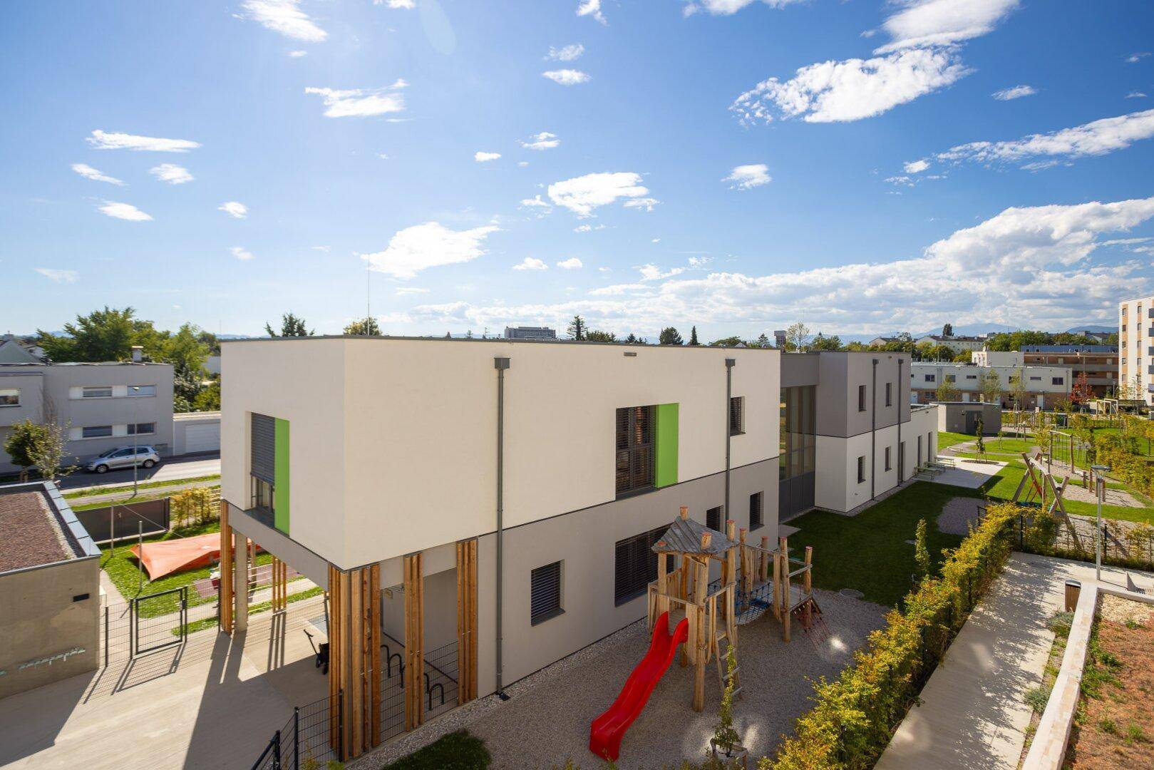 Moderne Gebäudefassade mit grünen Akzenten und angrenzendem Kinderspielplatz in der Wohnanlage.