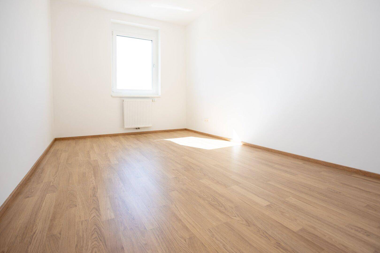 Helles Zimmer mit Laminatboden und einem Fenster, das den Raum mit Tageslicht erfüllt.