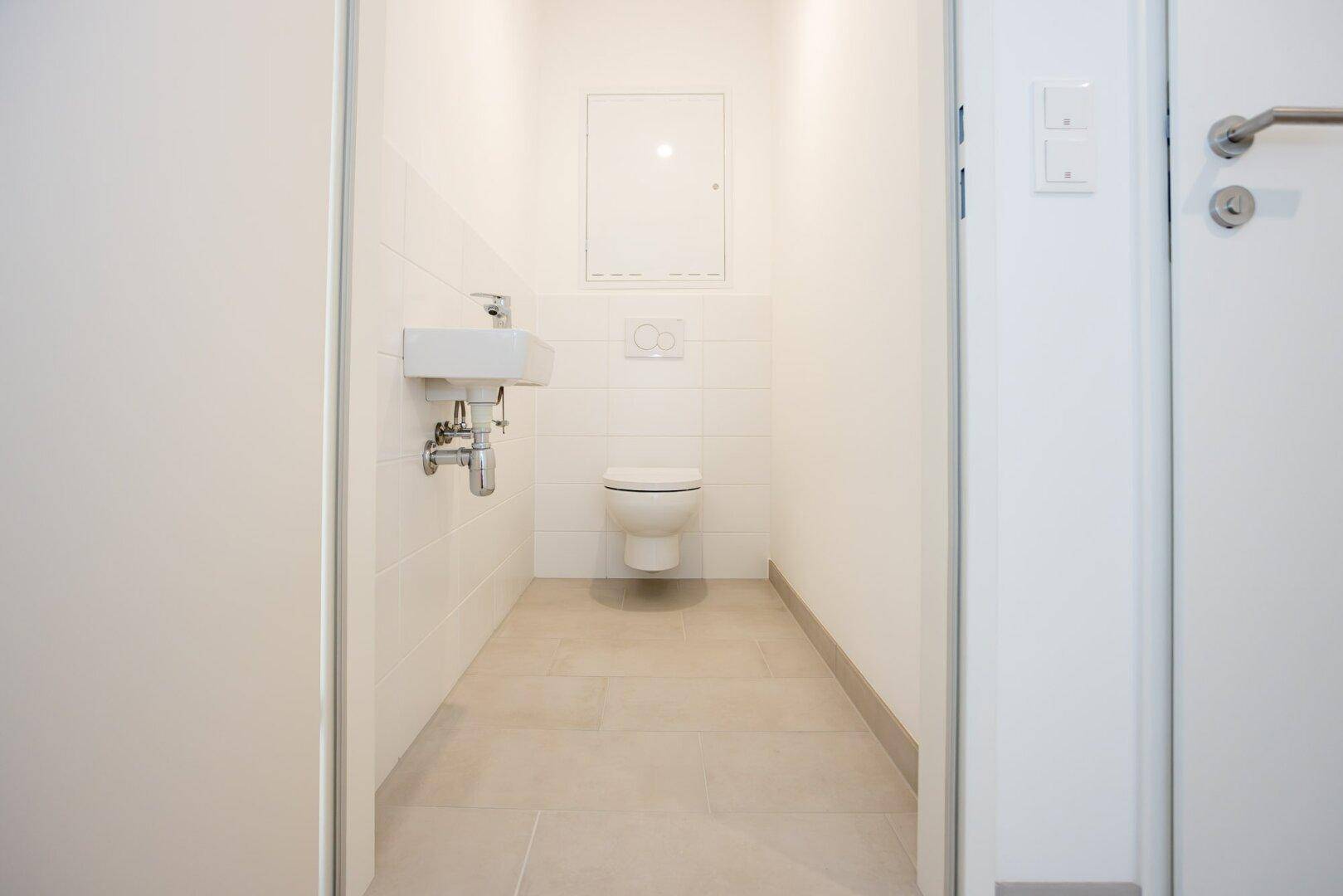 Separates Gäste-WC mit weißen Fliesen, wandhängendem WC und kleinem Handwaschbecken.