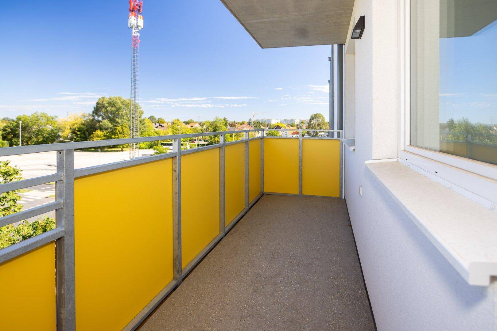 Heller Balkon mit gelben Brüstungselementen und Blick auf die Umgebung mit Mobilfunkmast.