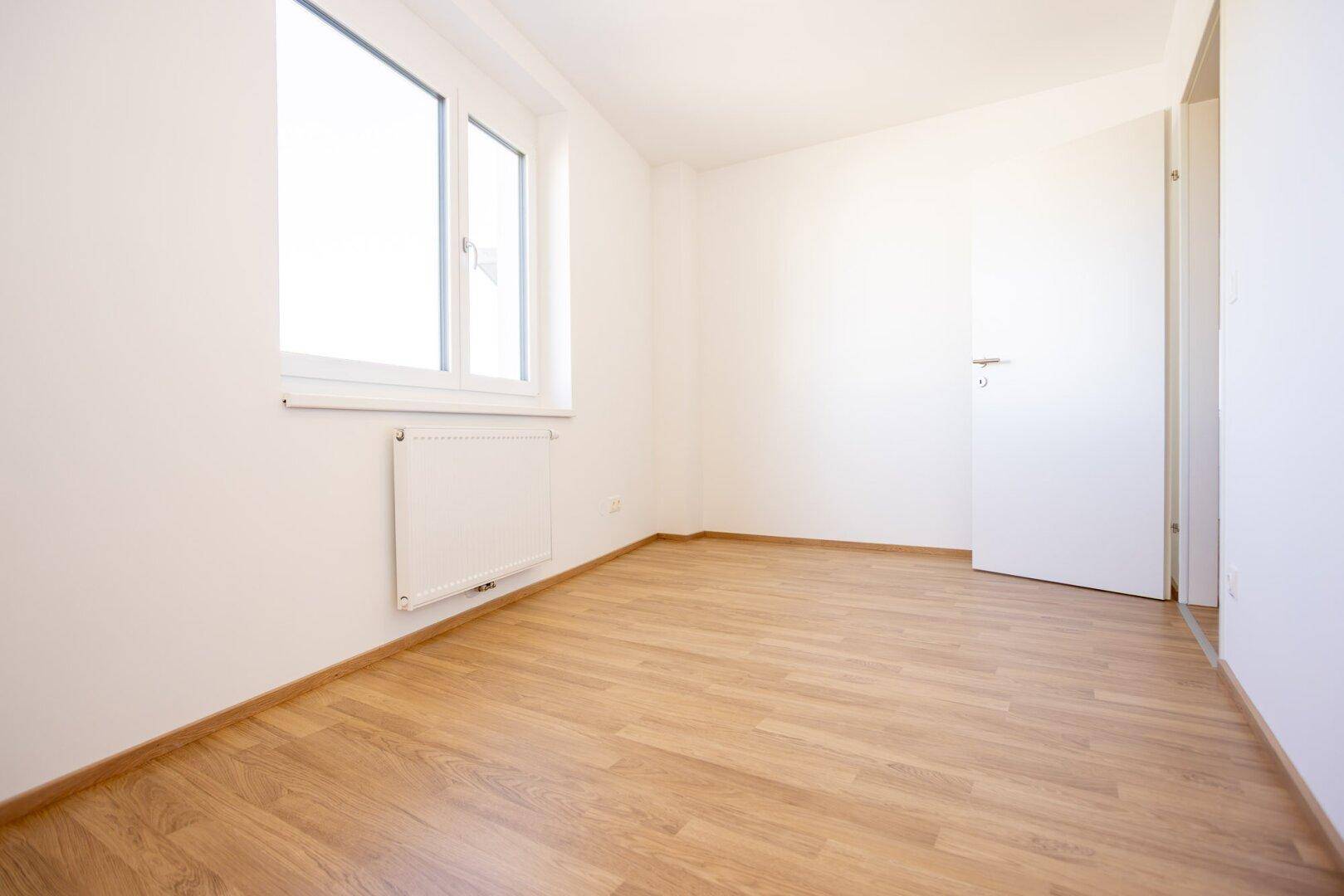 Helles Schlafzimmer mit Laminatboden, weißen Wänden und einem Fenster mit Heizkörper darunter.