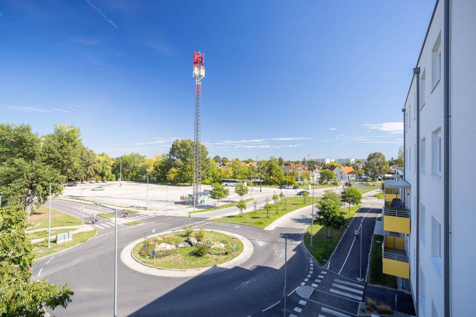 Weitläufige Ansicht einer städtischen Umgebung mit Kreisverkehr, Bäumen und einem Mobilfunkmast.